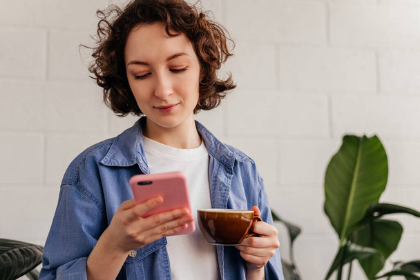 Młoda kobieta używająca telefonu komórkowego i pijąca kawę w kawiarni. Caicasian dziewczyna z kręconymi krótkimi włosami trzymając kubek wewnątrz. - Zdjęcie, obraz