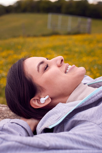 Woman Lying On Bench In Countryside Relaxing And Listening To Music Or Podcast On Wireless Earphones - Foto, imagen