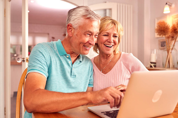 Senior Retired Couple Sitting On Sofa At Home Shopping Or Booking Holiday On Laptop - Fotoğraf, Görsel