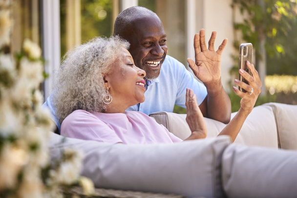 Senior Retired Couple Sitting Outside In Garden At Home Making Video Call On Mobile Phone - Foto, imagen