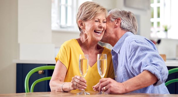 Kissing Retired Couple Celebrating With Glass Of Champagne At Home On Date Night Together - Foto, Imagen