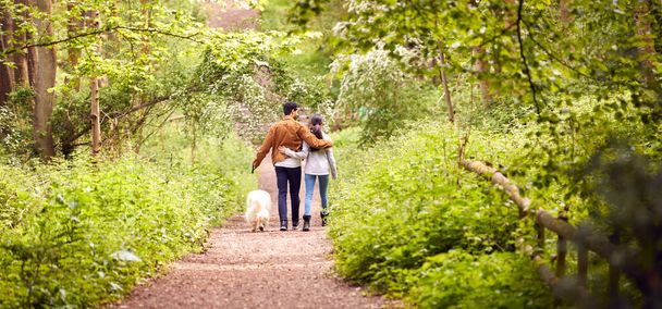Rear View Of Couple With Pet Dog Walking Along Path Through Trees In Countryside - 写真・画像