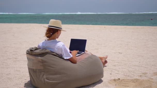 Erfolgreiche Geschäftsfrau mit gelbem Hut, die am Laptop am Meer arbeitet. Hübsche Dame tippt an einem Sommertag im Freien am Computer. Freiberufliches, Reise- und Urlaubskonzept. - Filmmaterial, Video