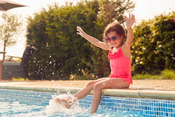 Girl Wearing Sunglasses Having Fun Splashing Sitting On Edge Of Swimming Pool On Summer Vacation - Foto, immagini