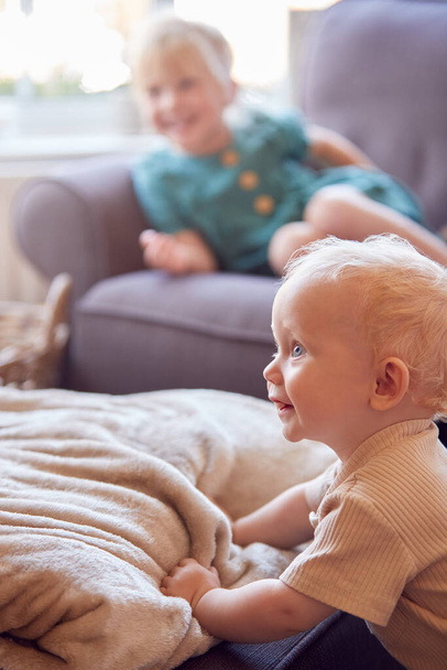 Young Girl Sitting On Sofa In Lounge At Home Watching TV As Baby Brother Plays In Foreground - Foto, Imagem