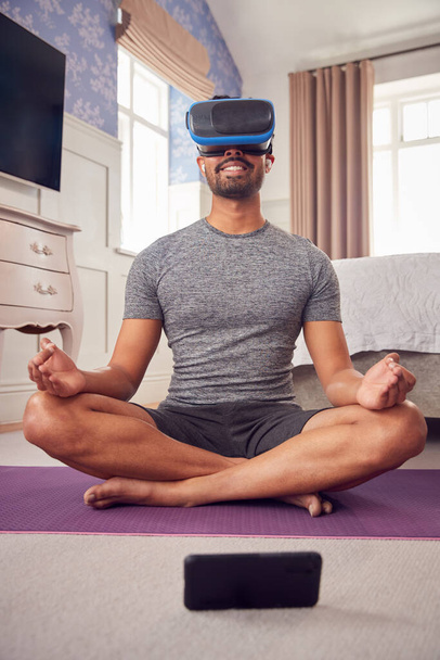 Man Wearing Virtual Reality Headset And Earphones Sitting On Yoga Mat In Bedroom At Home - Valokuva, kuva