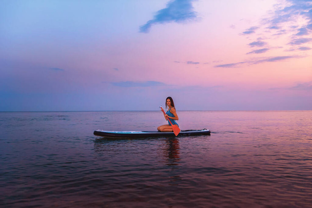 A young smiling woman poses sitting on a sup board with paddle. Sunset in the background. Copy space. Concept of sup boarding. - Фото, изображение
