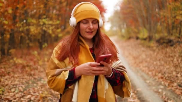 Aktiivinen valkoihoinen nainen kävelee yhdessä kuulokkeiden kanssa matkapuhelimella nauttii luonnosta kaupungin syyspuistossa. 5G teknologialaite. - Materiaali, video