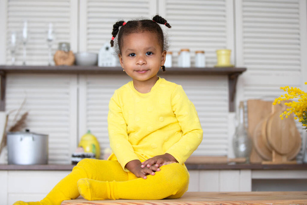 На кухні сидить маленька афроамериканська дівчинка в жовтій сукні з кучерявими хвостиками.. - Фото, зображення