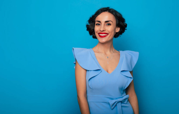 Close up portrait of gorgeous elegant brunette woman with wavy hairstyle and bright toothy smile while she posing in dress on blue background - Φωτογραφία, εικόνα