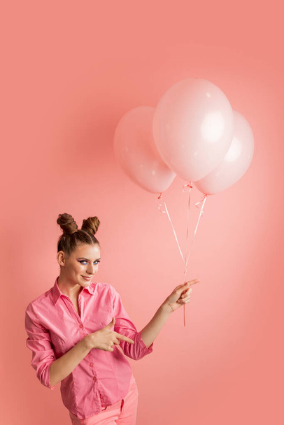 Porträt einer jungen Frau mit zwei Dutt-Frisuren und aufblasbaren Luftballons auf rosa Hintergrund. Mädchen zeigt mit dem Finger auf etwas nach rechts - Foto, Bild