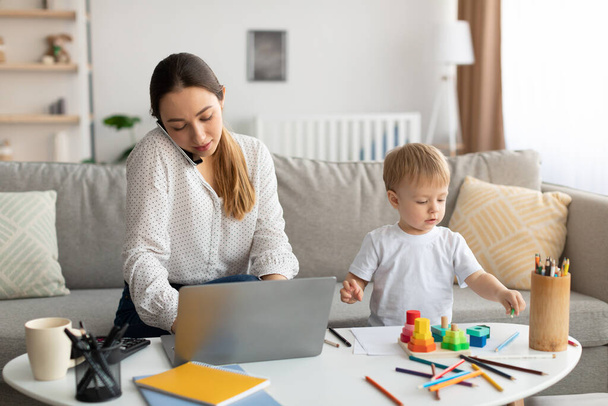 Bebekle çalışıyorum. Küçük oğlu yakınlarda oynarken, genç bir anne cep telefonuyla konuşuyor ve dizüstü bilgisayarda yazıyor. - Fotoğraf, Görsel