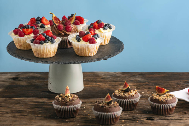 Vanilla and chocolate muffins with fresh berries on blue background. Confectionery on stand - Photo, Image