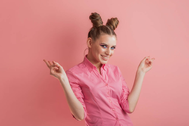 Joven chica sonriente en ropa rosa con peinado de moño sobre fondo rosa. Humor rosa - Foto, Imagen