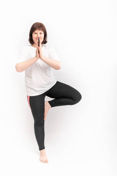 Young plump caucasian woman practices yoga. Portrait of girl in tree pose on white background. Vertical frame. - Photo, Image