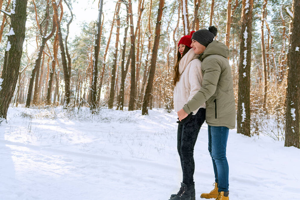 Joven pareja feliz están de pie y abrazándose en el día soleado en el parque cubierto de nieve. Hombres y mujeres en un parque invernal nevado. Vista lateral. - Foto, Imagen