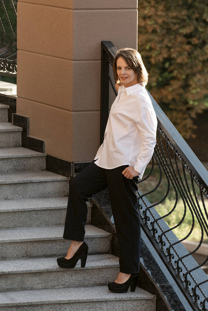 Stylish woman poses on steps with forged railings. Successful woman. Girl is dressed in classic white shirt and black pants. - Zdjęcie, obraz