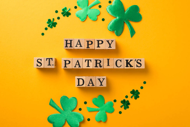 Top view photo of circle composition wooden cubes with words happy st patricks day surrounded by green trefoils and confetti on isolated yellow background - Photo, Image