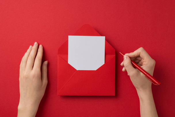 First person top view photo of st valentine's day decorations girl's hands holding pen over paper sheet in open red envelope on isolated red background with blank space - Фото, зображення