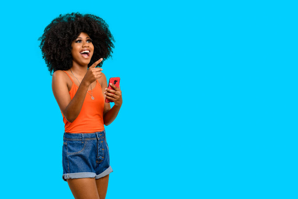 mujer joven con el pelo afro positivamente sorprendido apunta a un espacio blanco, ella sostiene un teléfono inteligente en una mano, aislado sobre fondo azul. - Foto, imagen