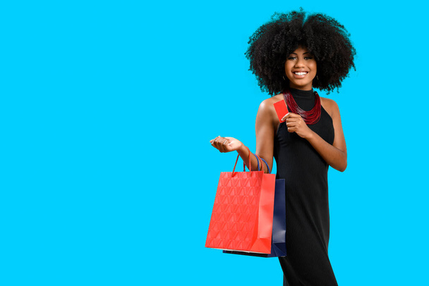 young woman with afro hair smiling happy holding a card and several shopping bags, in a long black dress, isolated on gray background. - 写真・画像
