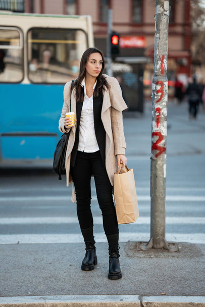 Hermosa mujer adulta joven con bolsa de compras se encuentra en un cruce peatonal esperando una luz verde para cruzar la calle. Ella es seria y mirando a un lado. - Foto, Imagen