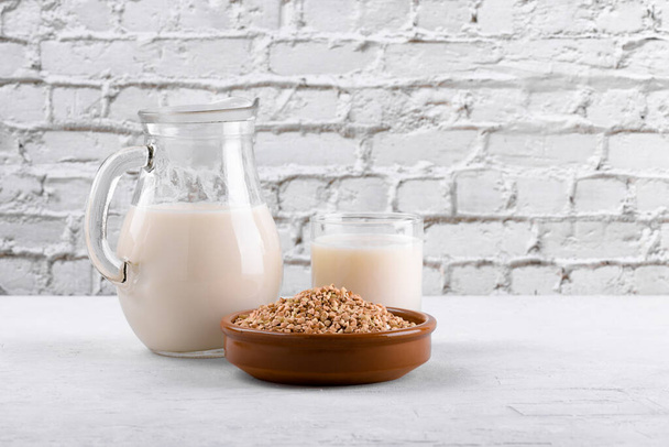 Vegan non ημερολόγιο φαγόπυρο γάλα σε κανάτα και γυαλί με βουβάλια φαγόπυρου ένα μπολ σε ένα λευκό πέτρινο τραπέζι. Vegan φαγόπυρο ποτό είναι φυτική βάση εναλλακτικό γάλα. Χορτοφάγα υγιεινό προϊόν γάλακτος σε στάμνα - Φωτογραφία, εικόνα