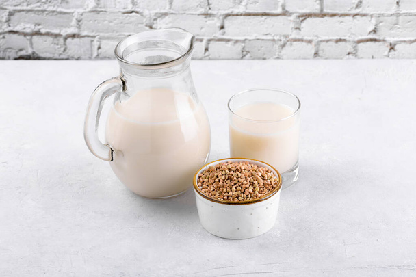 Vegane Buchweizenmilch in Krug und Glas mit Buchweizengrütze in einer Schüssel auf einem Tisch aus weißem Stein. Veganes Buchweizengetränk ist pflanzliche Alternativmilch. Gemüse gesundes Milchprodukt im Krug - Foto, Bild