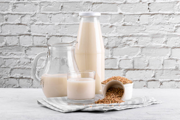Vegan non diary buckwheat milk in bottle, in jug and glass with buckwheat groats in a bowl on white stone table. Vegan buckwheat drink is plant based alternative milk. Veggies healthy milk product - 写真・画像