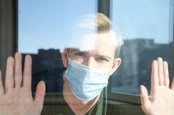 Hand of caucasian man in medical mask on glass of window on self-isolation during an epidemic. Blurry, defocus, light reflection. Man wants to leave the room and go outside. - Photo, Image