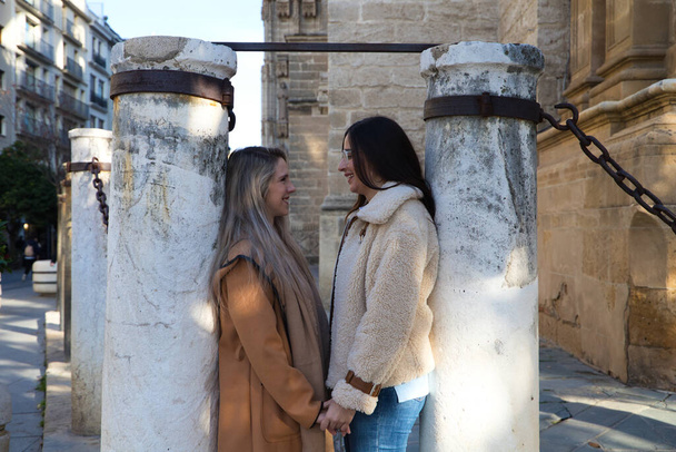 lesbisches Paar, das sich an Säulen lehnt, die die Kathedrale im gotischen Stil umgeben. Die Frauen sehen einander verliebt und glücklich an. Konzept der homosexuellen und lesbischen Ehe. - Foto, Bild