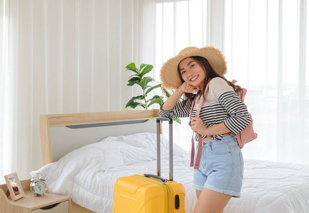 Yaz kıyafetleri, sırt çantası ve hasır şapkalı genç Asyalı kadın yatak odasında sarı bavulla duruyordu. Tatil gezisine hazırım. - Fotoğraf, Görsel