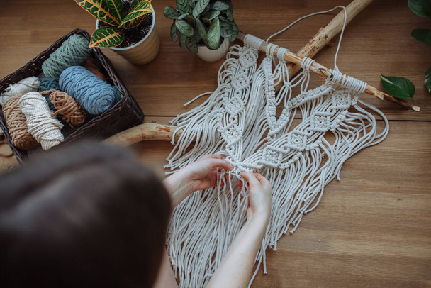 A brunette girl weaves a white panel using macrame technique on a wooden background. Nearby is a basket of macrame threads and houseplants. top view - Foto, afbeelding