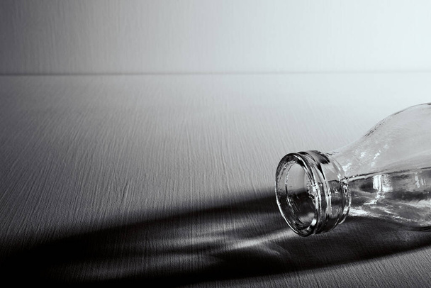 An empty glass bottle lies on the table. Copy space. Graphic still life with light and shadow in black and white colors. Concept of recyclable materials or alcoholism problems. Depressive condition. - Photo, Image