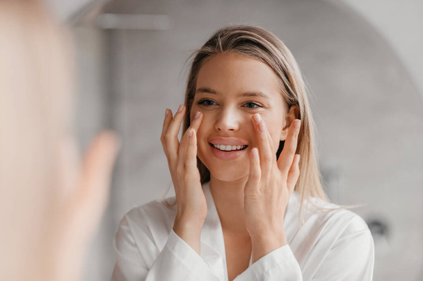 Jolie dame faisant massage lifting du visage dans la salle de bain, regardant le miroir et souriant, en utilisant crème hydratante visage - Photo, image