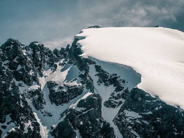 Jasny alpejski krajobraz z pokrytym śniegiem szczytem górskim i czarnymi skałami w świetle słonecznym. Alpejski minimalizm ze śnieżną górą na błękitnym niebie. Minimalny widok na szczyt śniegu na bardzo dużej wysokości. - Zdjęcie, obraz