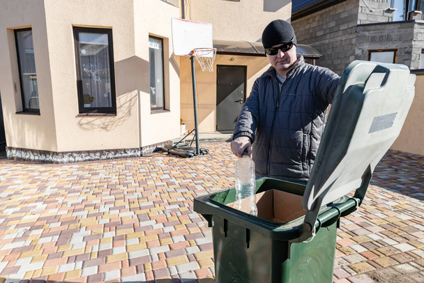 Ein Mann mittleren Alters wirft Plastikflaschen in einen Mülleimer im Hof eines Privathauses - Foto, Bild