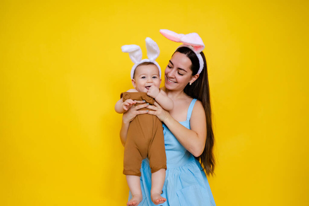 Μαμά και μωρό με αυτιά κουνελιού. Γονιός και παιδί παίζουν σε κίτρινο φόντο. Οικογένεια γιορτάζει το Πάσχα. Το χαριτωμένο αγοράκι φοράει αυτιά κουνελιού.. - Φωτογραφία, εικόνα