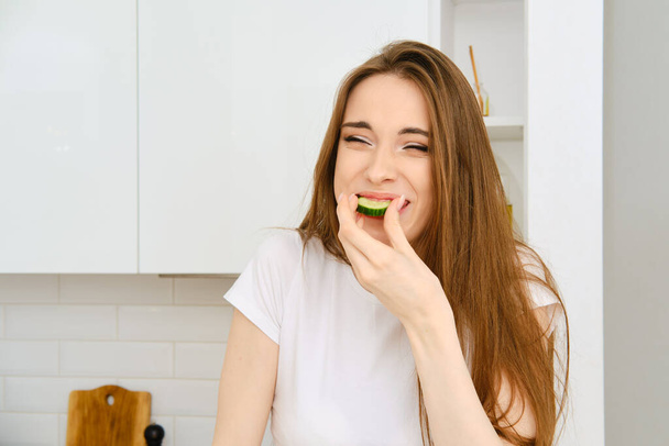 Funny young joyful woman in white t-shirt bites slice of cucumber, squints and smiling. Happy female having fun while cooking in modern white kitchen at home - Φωτογραφία, εικόνα