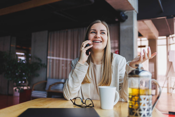 Young attractive female manager is talking on a mobile phone and smiling while sitting alone near a large window in a cafe in her free time and working on a computer. Happy woman is resting in a cafe. - 写真・画像