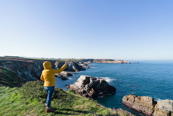 Mujer de abrigo amarillo tomando una selfie en un paisaje costero natural con mar y cielo azul. Copiar espacio. Fotografía horizontal. - Foto, imagen