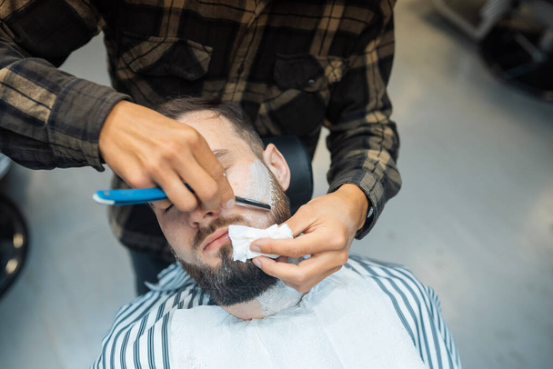 Young bearded man getting shaved by hairdresser at barbershop - Fotó, kép