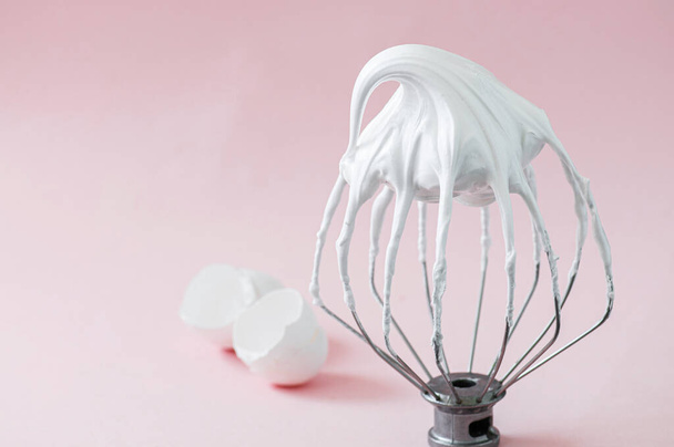 Whipped egg whites - beaten italian meringue on a wire whisk and egg shells on pink background.  - Foto, afbeelding