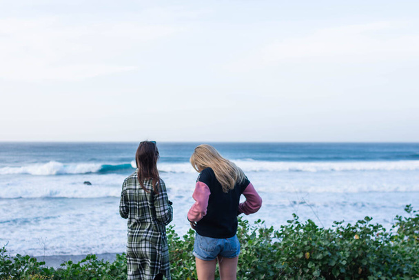 Una chica de pelo oscuro y una rubia de la espalda miran al mar en un día de fuertes olas en la playa - Foto, Imagen