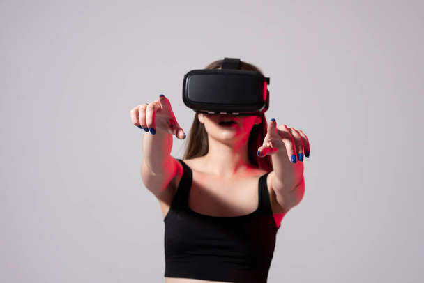 Brunette woman in a black t-shirt and VR headset looking up and trying to touch objects in virtual reality with both hands. Future technology concept. - Photo, image