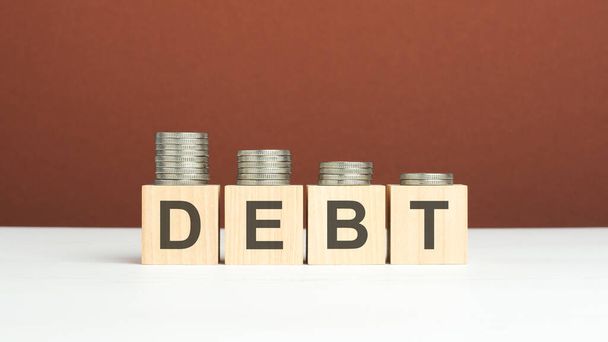 Schuldentext auf Holzklötzen mit Münzen auf braunem Hintergrund, Geschäftskonzept - Foto, Bild