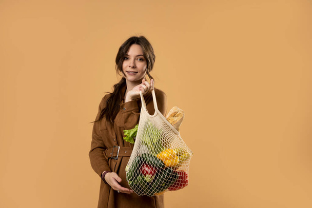Krásná atraktivní šťastná brunetka žena drží mesh potraviny taška se zeleninou. Koncept bez plastu. Bez odpadu, bez plastu. Ekologický koncept. Udržitelný životní styl. - Fotografie, Obrázek