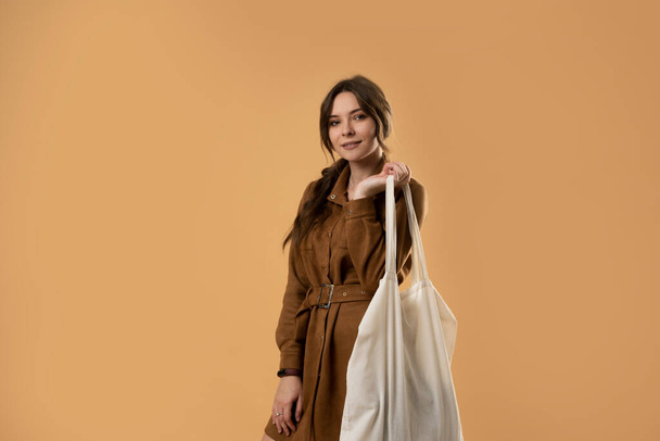Young attractive beautiful sexy brunette woman holding reusable mesh bag with organic groceries over orange background. Zero waste, plastic free concept. Eco lifestyle. Conscious consumption. - 写真・画像