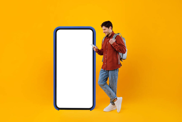 Traveler Guy χρησιμοποιώντας τηλέφωνο στέκεται κοντά στο μεγάλο κινητό τηλέφωνο, κίτρινο φόντο - Φωτογραφία, εικόνα