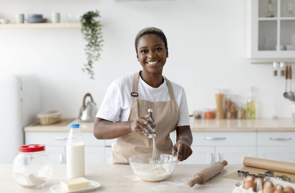 Lächelnd hübsche afrikanisch-amerikanische Köchin in Schürze, die Teig im leichten minimalistischen Kücheninterieur zubereitet - Foto, Bild